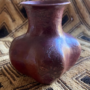 May include: A handcrafted, burgundy-colored vase with a textured surface. The vase features a floral design carved into the front. The vase has a wide, rounded body and a flared top. The vase is likely made of metal.