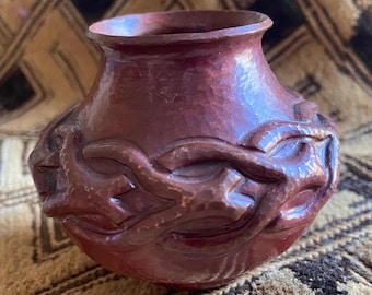 May include: A handcrafted, reddish-brown ceramic vase with a rounded body and a slightly flared rim. The vase features a carved, repeating design around its midsection, adding texture and visual interest. The piece has a rustic, artisanal aesthetic.
