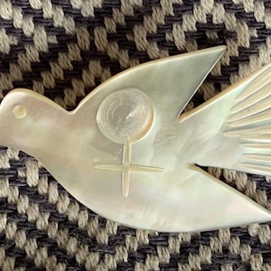 May include: A pearlescent dove-shaped brooch with a cross and circle design. The bird has detailed wing feathers and a smooth body. The brooch is set against a textured, neutral-toned background.