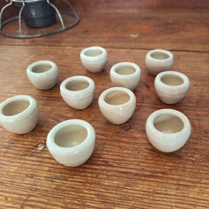 Könnte beinhalten: Eine Ansammlung von neun kleinen, runden, cremeweißen Keramikgefäßen mit einer glatten, glänzenden Oberfläche. Jedes Gefäß hat eine weite Öffnung und einen abgerundeten Boden und ist verstreut angeordnet, um die einheitliche Form zu zeigen.