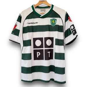 Puede incluir: Camiseta de fútbol a rayas blancas y verdes con el escudo del Sporting Portugal en el pecho. La camiseta tiene cuello en V y el logo de Reebok. Las mangas tienen ribete negro y las letras "PT" están en la parte delantera.