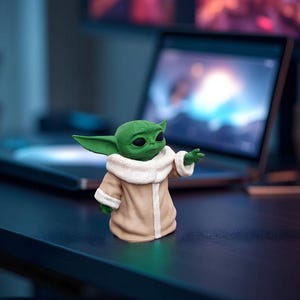 以下が含まれることがあります： 大きな耳を持つ緑色のキャラクターのフィギュア。白い襟のついたベージュのコートを着ています。フィギュアは暗い表面にあり、背景にはラップトップとモニターがあります。