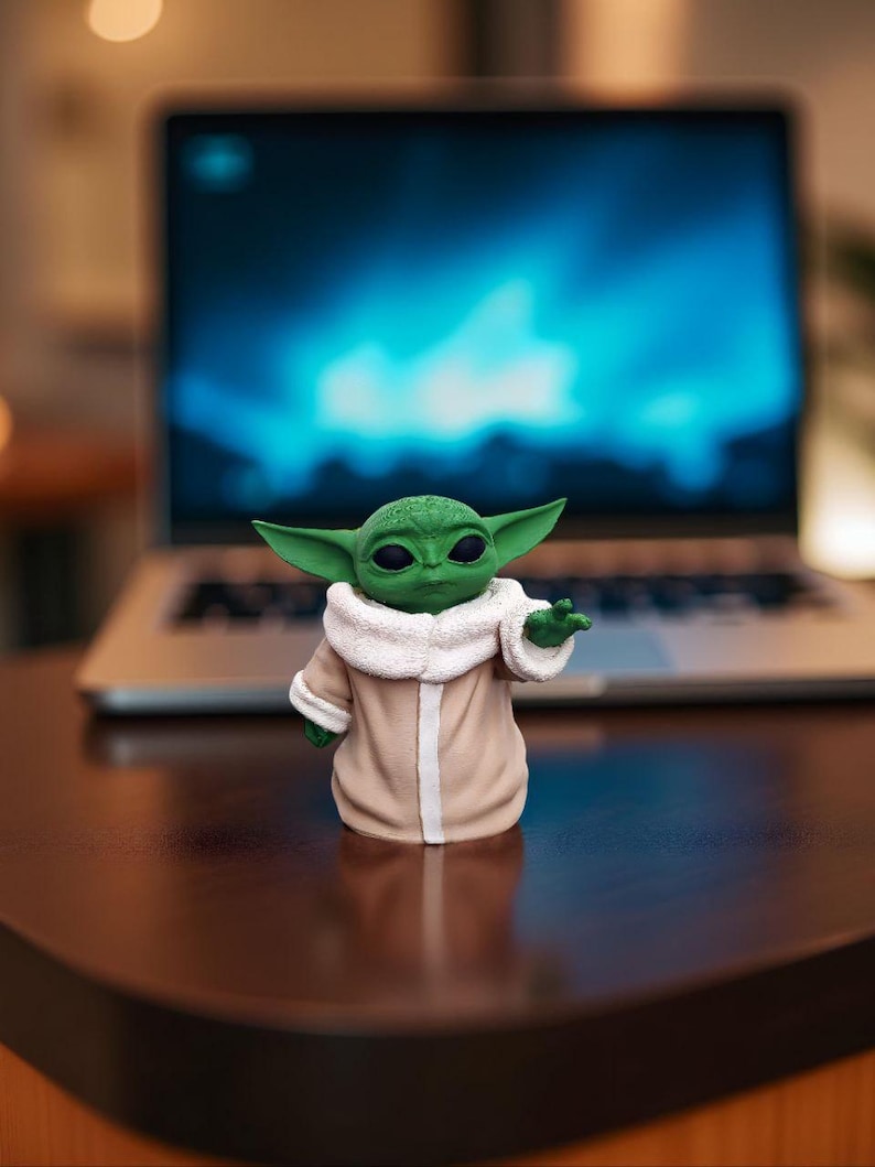 以下が含まれることがあります： 大きな耳を持つ小さな緑色の肌のフィギュアで、白い毛皮のような襟が付いたベージュ色のローブを着ています。フィギュアは片手を上げてポーズをとっています。青い画面のラップトップが背景にあります。