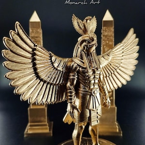Peut inclure: Une statue dorée d'une divinité égyptienne avec de grandes ailes, une coiffe et des détails complexes. Deux obélisques dorés flanquent la figure centrale. Le texte "Monarch Art" est visible dans le coin supérieur droit.