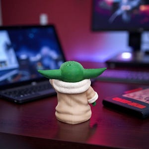 以下が含まれることがあります： 人気のSFフランチャイズのキャラクターの3Dプリントフィギュア。フィギュアは緑色の頭と大きな耳を持ち、白い襟のついたタン色のコートを着ています。フィギュアは濃い茶色の机の上にあります。