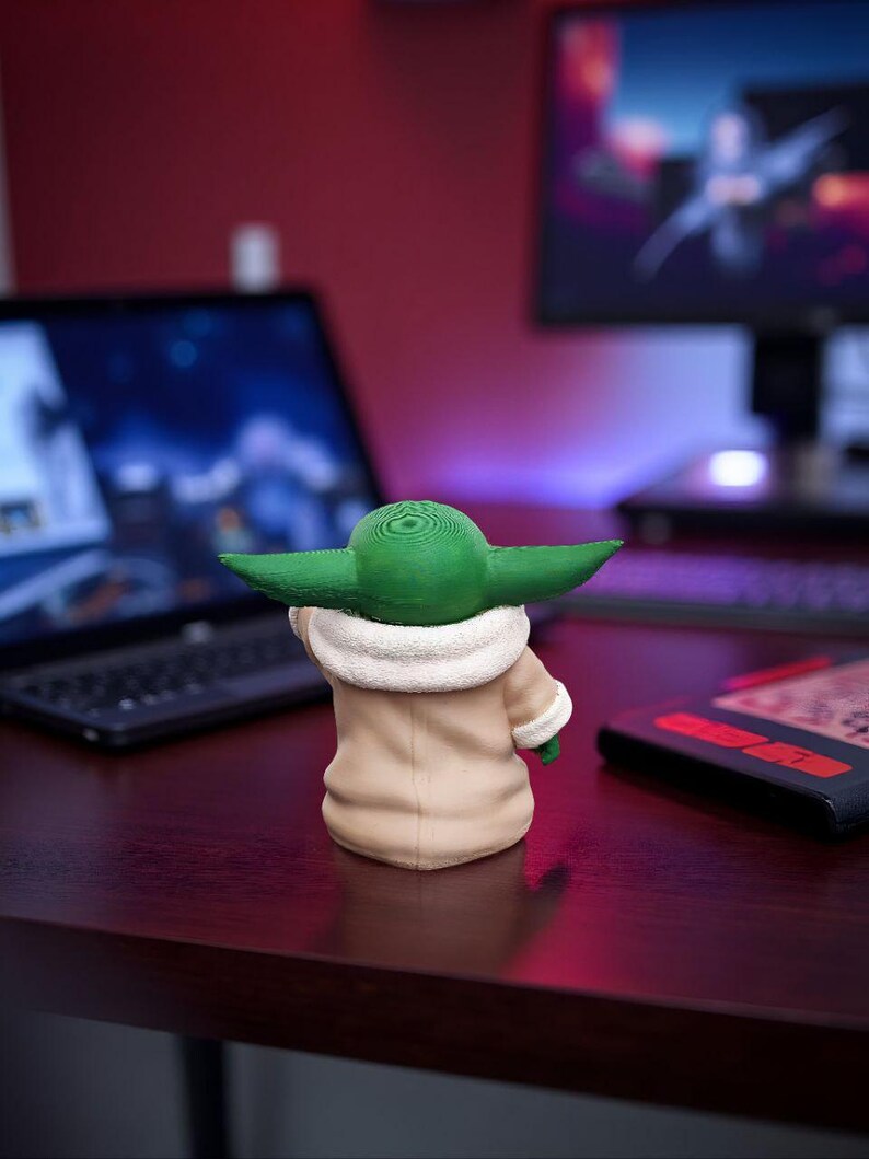 以下が含まれることがあります： 人気のSFフランチャイズのキャラクターの3Dプリントフィギュア。フィギュアは緑色の頭と大きな耳を持ち、白い襟のついたタン色のコートを着ています。フィギュアは濃い茶色の机の上にあります。