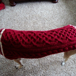 Op de afbeelding: Een rode gebreide hondentrui met witte strepen en een kabelbreipatroon.