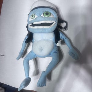 May include: A light blue plush character toy with a wide, toothy grin and green eyes. The figure wears a white pilot-style cap and a black vest with straps. It has long, thin limbs, a rounded belly, and dark blue spots on its arms and legs.