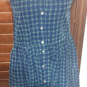 May include: A blue and green plaid dress with silver buttons. The sleeveless dress is displayed on a white mannequin. The dress has a button-down front and a gathered waist, creating a classic, vintage look.