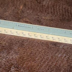 Puede incluir: Una regla de escala arquitectónica de doble cara en azul claro y blanco roto. La regla presenta medidas imperiales y métricas, con marcas claras para mediciones precisas. La escala está diseñada para dibujos arquitectónicos y de ingeniería.