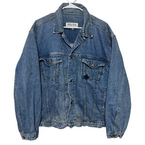 May include: A medium-wash blue denim jacket featuring a classic collar, long sleeves with buttoned cuffs, and a front button closure. The garment includes two chest pockets with buttoned flaps and a white label reading BUGLE BOY at the neck.