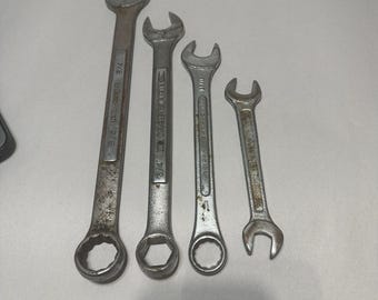 May include: Four metal wrenches of varying sizes are arranged on a white surface. The wrenches have a combination of open-end and box-end designs. The largest wrench is marked with "9/16" and "3/4". The tools show signs of wear.