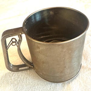 Puede incluir: Un tamizador de harina de metal vintage con asa. El tamizador es cilíndrico con una parte superior ligeramente ensanchada y un asa de alambre. El metal tiene un aspecto opaco y envejecido, lo que sugiere que es una herramienta de cocina antigua. El asa tiene un diseño decorativo.