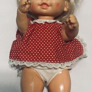 Puede incluir: Muñeca de bebé vintage con cabello rubio y ojos azules. La muñeca viste un vestido rojo con lunares blancos y un ribete de encaje blanco. La muñeca es de plástico. Las mejillas de la muñeca son rosadas.