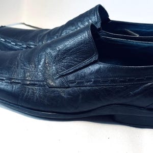 May include: A pair of black leather slip-on shoes. The shoes have a square toe design with white stitching along the top and sides. The leather appears textured and worn. The shoes are on a white surface.
