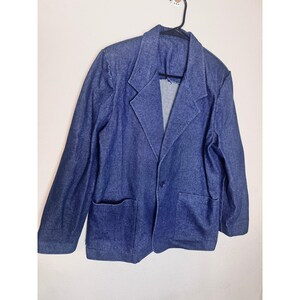 May include: A dark blue denim blazer featuring a notched lapel, a single-button front closure, and two large patch pockets on the lower front. The jacket has long sleeves and is displayed on a black plastic hanger against a plain wall.