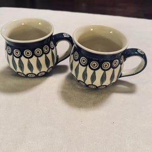 May include: Two ceramic mugs with a blue and white floral pattern. The mugs have a rounded shape with a handle and a cream-colored interior. The design includes blue circles with red centers and green leaf-like shapes.