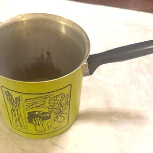 May include: A lime green metal saucepan with a black handle and a silver interior. The exterior features a black line art graphic depicting various vegetables including asparagus, broccoli, and radishes, with the text labels printed below.
