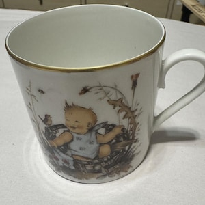 Può includere: Tazza in ceramica bianca con bordo dorato e manico curvo. La tazza presenta un'illustrazione stravagante di un bambino in un cesto circondato da piante e un uccellino. Il design è in colori tenui e smorzati.