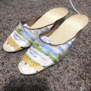 Puede incluir: Un par de zapatillas de estilo vintage con un diseño de paisaje. Las zapatillas presentan una base color crema con una escena pintada en tonos azules, verdes y amarillos. Los bordes están ribeteados en marrón y las plantillas son de color beige claro.