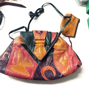 May include: A colorful, patchwork-style handbag with a shoulder strap. The bag features a variety of textures and colors, including orange, pink, green, and purple. The bag has a flap closure with a metal clasp and a small matching pouch.