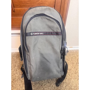 May include: An olive green backpack featuring a black horizontal stripe with the white text "tamrac" and a thin red line. The bag includes black zips, black adjustable straps, and a small silver padlock attached to the side zip pull.