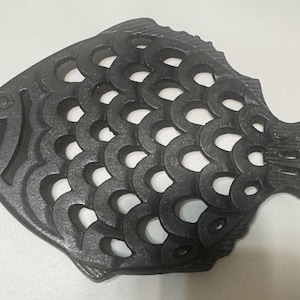 Può includere: Un sottopentola in ghisa grigio scuro a forma di pesce. Il pesce presenta un design dettagliato con cerchi aperti che formano le squame. Il sottopentola è progettato per proteggere le superfici dalle pentole calde.