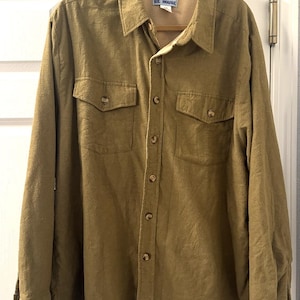 May include: Olive green button-down shirt with two chest pockets. The shirt has a collared neckline and long sleeves. The tag reads "HARARD'S ICE HOUSE". The shirt is hanging on a wooden hanger.