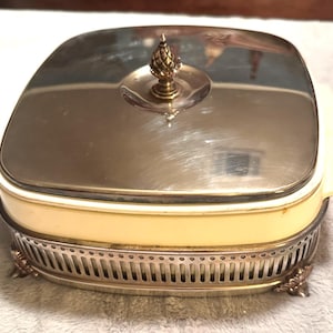 Könnte beinhalten: Eine Vintage-Servierschale mit silberfarbenem Deckel und dekorativer goldfarbener Verzierung. Die Schale hat einen cremefarbenen Boden und einen silberfarbenen Metall-Filigranboden mit dekorativen Füßen. Der Deckel ist quadratisch.