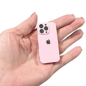 May include: A miniature light pink smartphone replica featuring a dark gray camera module with three circular lenses and a small dark gray apple logo centered on the back. The device has a smooth, matte finish and rounded corners, mimicking a modern mobile phone design in a small scale.
