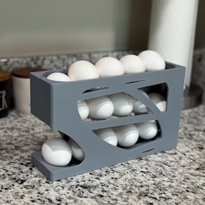 May include: A gray, two-tiered egg holder filled with white eggs. The holder has a modern design with cut-out shapes on the sides. The eggs are visible in the open compartments, providing a clear view of the contents.
