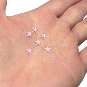 May include: A close-up of a hand holding several small, light pink star-shaped sprinkles. The sprinkles are scattered across the palm, with a few visible in the center and towards the edges of the hand.