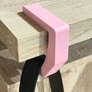 May include: A light pink purse hanger is attached to the edge of a wooden table. The hanger is a C-shaped design, with a flat top and a curved bottom. A black purse strap hangs from the hanger.