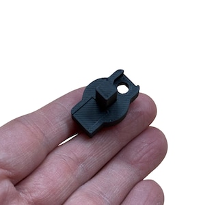 May include: A small, black, plastic component with a square protrusion and a circular hole. The object has a unique shape with angled edges and appears to be 3D-printed. The component is held in a hand against a white background.