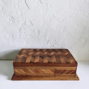 May include: A rectangular wooden box featuring a prominent herringbone pattern on the lid and sides. The box is constructed from various shades of brown wood, showcasing natural grain variations and a smooth finish on the exterior surfaces.