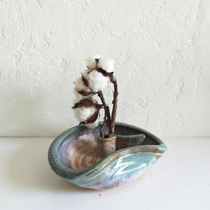 May include: A ceramic decorative bowl with a unique shape and a blend of blue, purple, and brown glaze. It holds a small vase with cotton stems, creating a natural and artistic display. The bowl's design is organic and visually appealing.