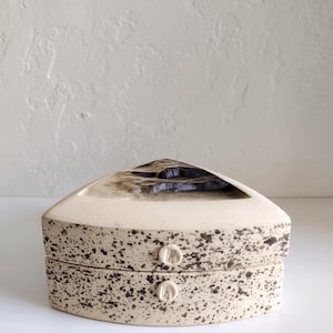 May include: A speckled ceramic box with a triangular lid and two small, round handles. The box is off-white with dark brown speckles and a dark interior. The lid has a unique, open design.