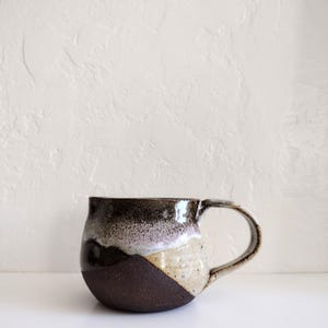 May include: A handcrafted ceramic mug with a unique design. The mug features a dark brown base, transitioning to a speckled beige and then a dark brown and black rim. It has a curved handle and a smooth, glossy finish.
