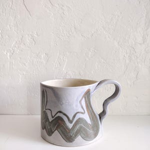 May include: A ceramic mug with a light blue glaze and a gray handle. The mug features a wavy, abstract design in shades of gray and brown. The interior of the mug is a light cream color. The mug is on a white surface.