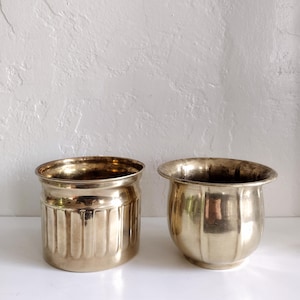 May include: Two brass planters with a vintage aesthetic. One is cylindrical with vertical grooves, the other is rounded with a flared rim. Both have a warm, metallic sheen, perfect for displaying small plants or decorative items.