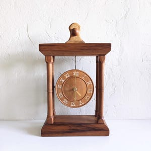 May include: A wooden mantel clock featuring a rectangular frame with four pillars and a central circular dial. The dial displays white Roman numerals and gold-colored hands. A small decorative wooden finial sits atop the clock structure.
