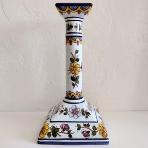 May include: White ceramic candlestick with a square base and floral design. The candlestick features yellow, pink, and blue flowers with green leaves. The edges are trimmed with blue and yellow accents.