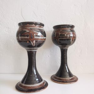 May include: Two handmade ceramic goblets. The goblets are dark brown with a bulbous top and a long stem. The top of each goblet has a brown and white geometric design. The base of each goblet is round and has a brown and white stripe.