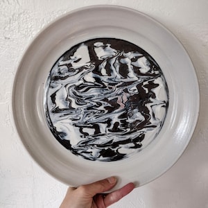 May include: A white ceramic plate with a black and white marbled design in the center. The plate has a raised rim.