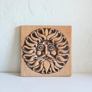 May include: A square terracotta-colored ceramic tile featuring a circular relief carving of a sun face with a bearded human expression. The design includes stylized flame-like rays radiating outward, with dark brown accents highlighting the recessed details.