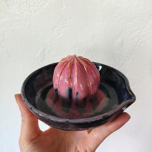 May include: A pink and black ceramic citrus juicer with a ridged top and a small spout. The juicer is sitting on a black and pink ceramic dish.