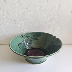 May include: A wide, circular ceramic bowl with a glossy sage green glaze and a dark, deep purple interior base. The rim features a small notch on one side and two small circular holes on the opposite side, with dark brown abstract brushwork.