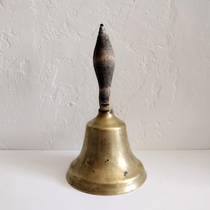 May include: A vintage brass bell with a wooden handle. The bell has a classic shape with a flared base and a rounded top. The handle is dark brown and textured, contrasting with the shiny brass. The bell is likely used for signaling or decoration.