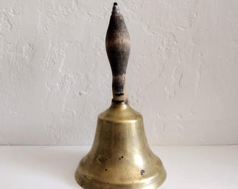 May include: A vintage brass bell with a wooden handle. The bell has a classic shape with a flared base and a rounded top. The handle is dark brown and textured, contrasting with the shiny brass. The bell is likely used for signaling or decoration.