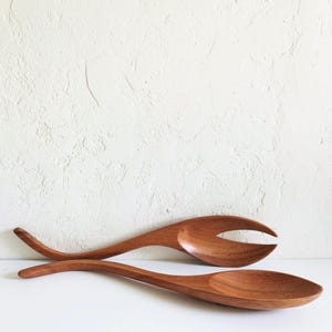 May include: Two wooden salad servers. One server is a spoon, and the other has a fork-like design. Both are made of a warm brown wood and have long, curved handles. The servers are displayed on a white surface.